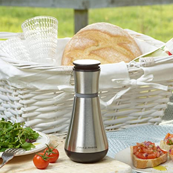 A Cole & Mason UK Henley Oil & Vinegar Pourer 180mm sits on a picnic table beside a stainless steel grinder, a white wicker bread basket, clear glasses, tomatoes, salad, and bruschetta with lush greenery in the background.
