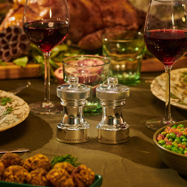A dinner table set with wine glasses, plates of food, and a pair of shiny metallic salt and pepper grinders in the center, surrounded by festive dishes including vegetables and a roast in the background.