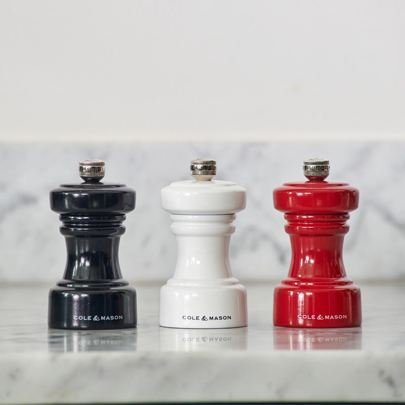 Three Cole & Mason mills—black, white, and red—stand side by side on a marble countertop in front of a light-colored marble backsplash.