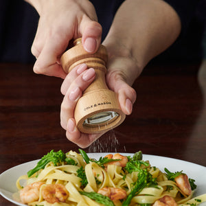 A person grinds fresh pepper onto a plate of pasta with shrimp and broccoli using a wooden pepper mill labeled Cole & Mason.