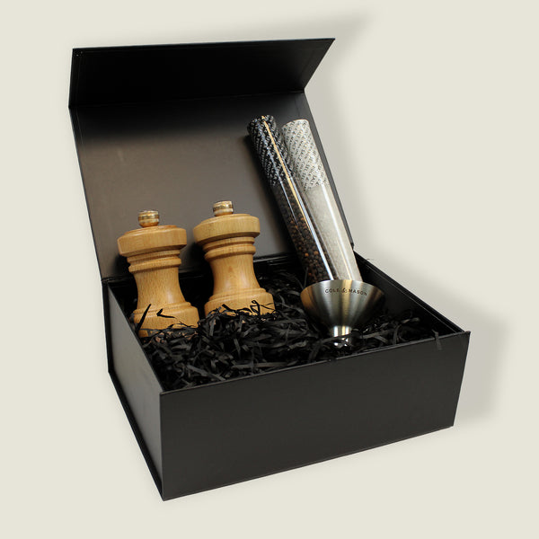 The Cole & Mason Hoxton Natural Beech Mill Christmas Gift Hamper features two wooden mills, two tubes of black peppercorns, and a stainless steel funnel in a black gift box with shredded paper—perfect for Christmas hamper boxes.