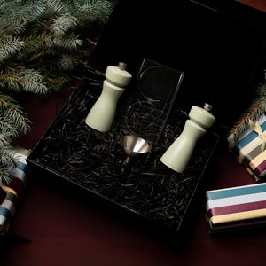 The Kenton Mills in Sage Christmas Gift Hamper by Cole & Mason includes two green grinders, a small metal funnel, and a clear accessory in black shredded paper, accented with pine branches and striped presents.