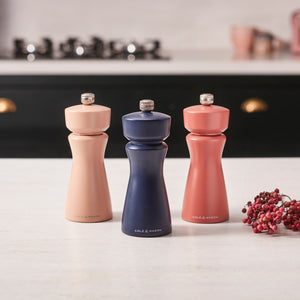 A Cole & Mason Kenton Salt & Pepper Mill in Persian Blue Salt stands on a kitchen counter alongside two other mills in peach and coral shades, all featuring ceramic grinders. Pink peppercorns are scattered nearby, and a stove is visible in the background.