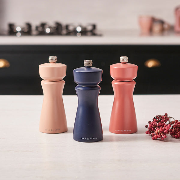 A Cole & Mason Kenton Salt & Pepper Mill in Persian Blue Salt stands on a kitchen counter alongside two other mills in peach and coral shades, all featuring ceramic grinders. Pink peppercorns are scattered nearby, and a stove is visible in the background.