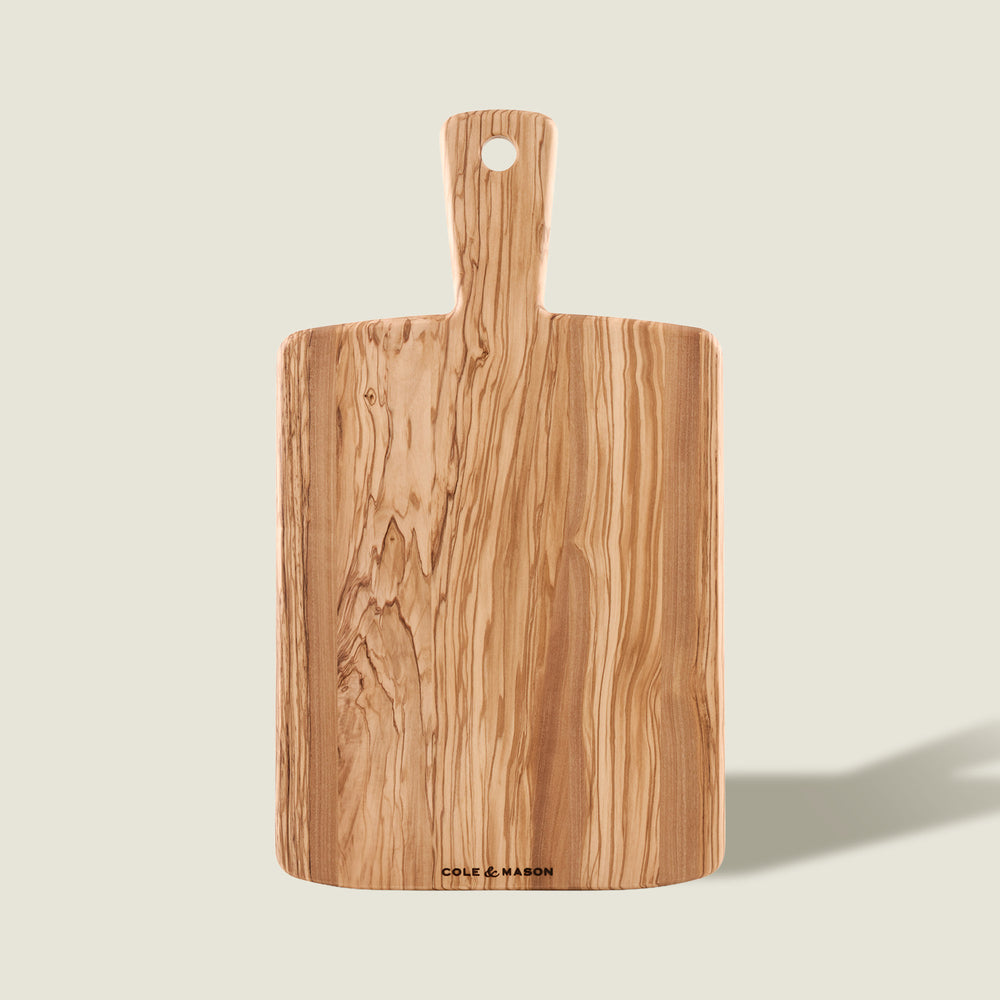 The Cole & Mason Portobello Olive Wood Serving Board, with a handle and hanging hole, showcases an elegant natural grain—ideal as a stylish antipasti board and shown upright on a light background.