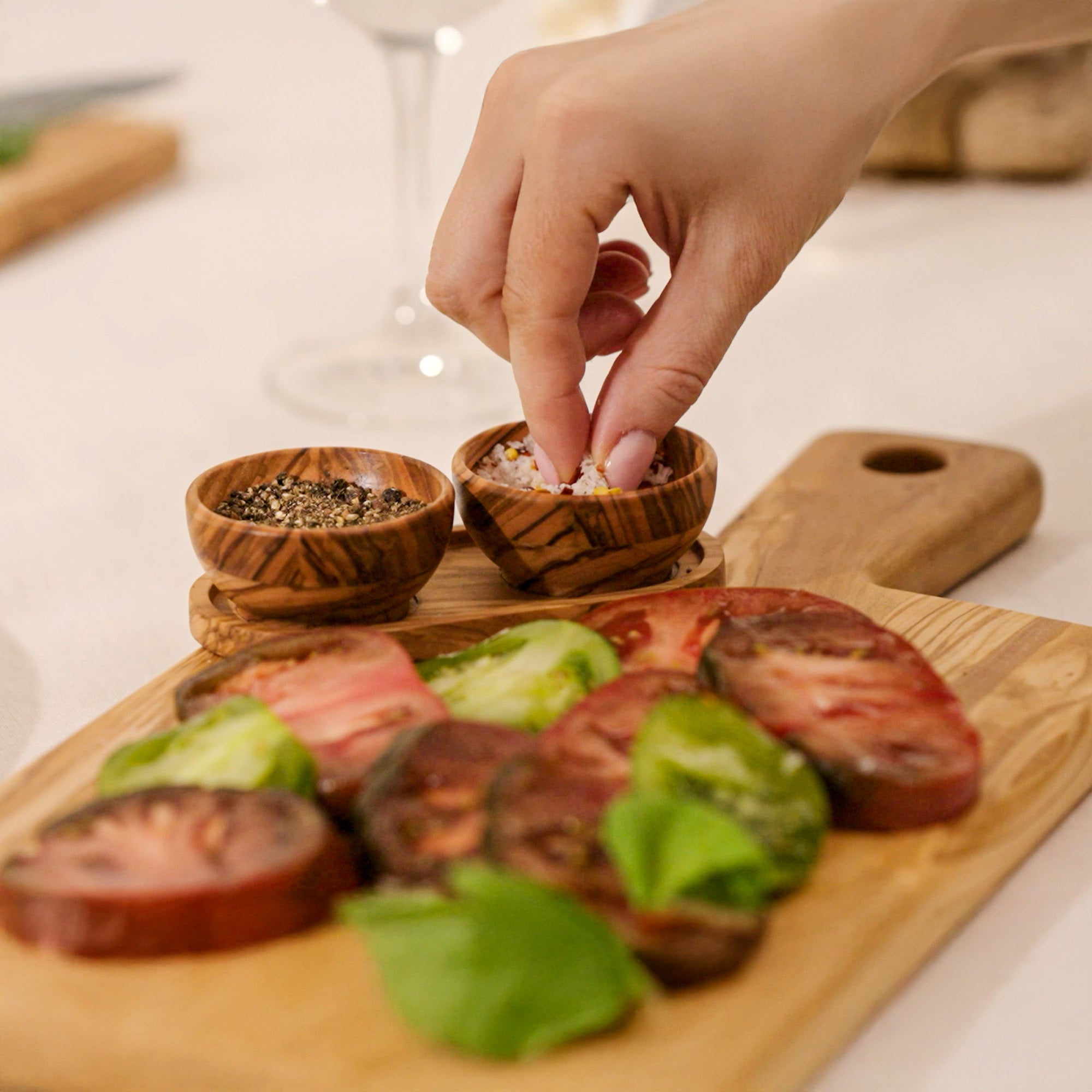 A hand sprinkles seasoning onto sliced heirloom tomatoes and basil, beautifully arranged on the Cole & Mason Portobello Olive Wood Serving Board & Pinch Pot Set. Nearby, pinch pots hold ground pepper for finishing touches.