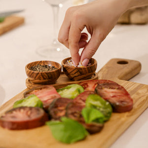 A hand sprinkles seasoning onto sliced heirloom tomatoes and basil, beautifully arranged on the Cole & Mason Portobello Olive Wood Serving Board & Pinch Pot Set. Nearby, pinch pots hold ground pepper for finishing touches.