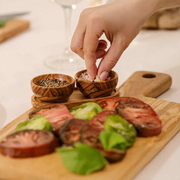 A hand sprinkles seasoning onto sliced heirloom tomatoes and basil, beautifully arranged on the Cole & Mason Portobello Olive Wood Serving Board & Pinch Pot Set. Nearby, pinch pots hold ground pepper for finishing touches.