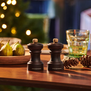 The Cole & Mason Hoxton Chocolate Wood Mill Christmas Gift Hamper, set on a wooden table with festive décor and a blurred Christmas tree in the background, is perfect for holiday gatherings or as a luxury treat in hamper boxes.