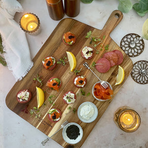 The Cole & Mason 2 Piece Barkway Board Bundle displays appetizers, lemon wedges, smoked salmon, salami, dips, caviar, and microgreens. Surrounded by candles on marble with a white cloth, it adds elegance to any gathering.