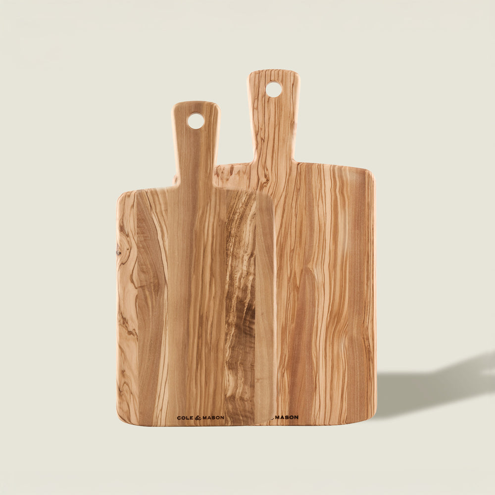 The Cole & Mason Portobello Olive Wood Serving Boards Set features two handled boards with visible grain, standing upright against a light beige background. The larger board is positioned behind the smaller; both have a hole at the end of the handle.