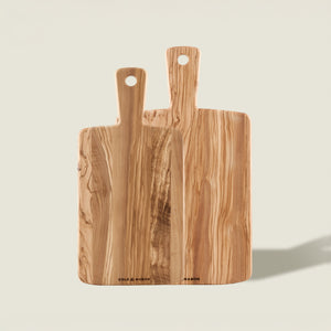 The Cole & Mason Portobello Olive Wood Serving Boards Set features two handled boards with visible grain, standing upright against a light beige background. The larger board is positioned behind the smaller; both have a hole at the end of the handle.