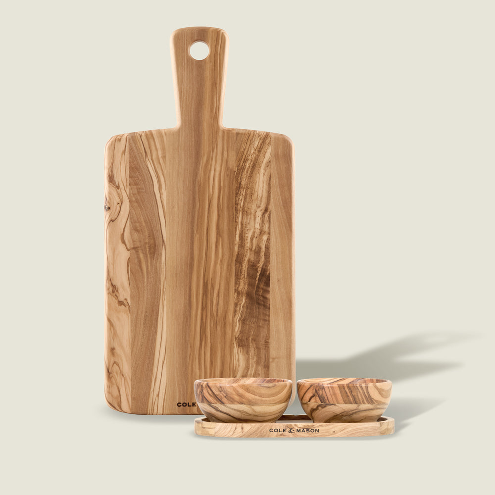 The Cole & Mason Portobello Olive Wood Serving Board & Pinch Pot Set features a handled board standing behind two small pinch pots and a tray, all displaying beautiful natural wood grain patterns on a light background.