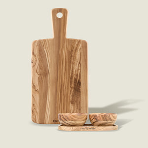 The Cole & Mason Portobello Olive Wood Serving Board & Pinch Pot Set features a handled board standing behind two small pinch pots and a tray, all displaying beautiful natural wood grain patterns on a light background.