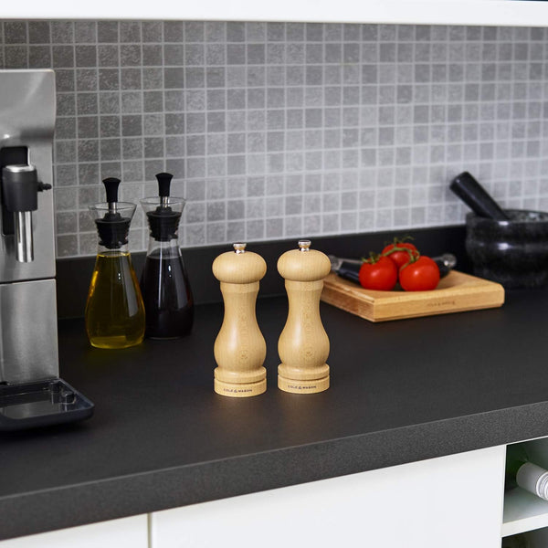A modern kitchen countertop with a coffee machine, Cole & Mason Beech Capstan Mill Christmas Gift Hamper, bottles of oil and vinegar, mortar and pestle, cutting board with tomatoes, and grey tiled backsplash.