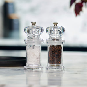 The Cole & Mason 505 Salt & Pepper Mills sit side by side on a marble counter, each filled with salt or pepper. Their clear acrylic design and adjustable settings stand out against the softly blurred background for a modern touch.