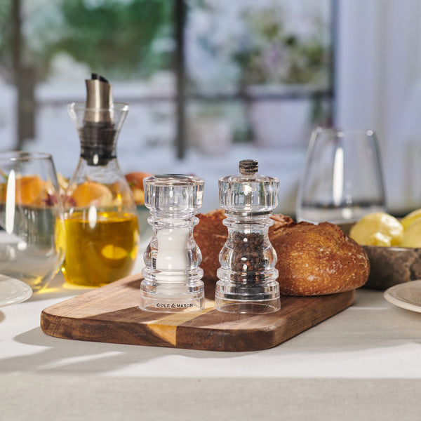 A wooden board displays the Cole & Mason London Acrylic Salt Shaker & Pepper Mill Set, surrounded by bread, butter, olive oil, and glasses, set near a bright window.