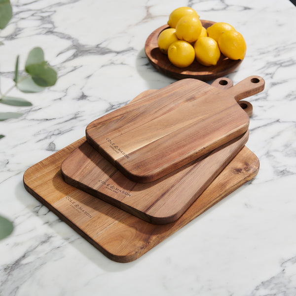 The 2 Piece Barkway Board Bundle by Cole & Mason, featuring boards of different sizes, is stacked on a marble countertop. Nearby, a small wooden bowl holds ripe yellow lemons with green leaves in the corner—an ideal kitchen essential.