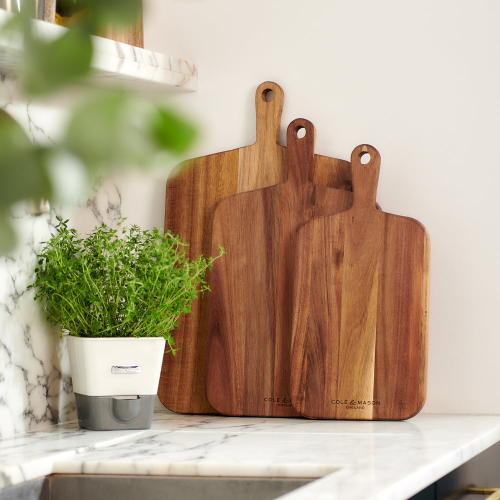 The Cole & Mason 3 Piece Barkway Board Bundle features three rectangular acacia chopping boards with handles, arranged overlapping to showcase their premium wood grain—each stamped with the brand name for a stylish kitchen essential.