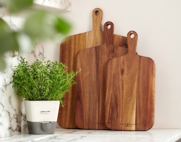 Two Cole & Mason Barkway Boards stand upright on a marble countertop beside a potted green herb. Crafted from premium acacia wood, this 2 Piece Board Bundle is an essential kitchen addition for any home chef.