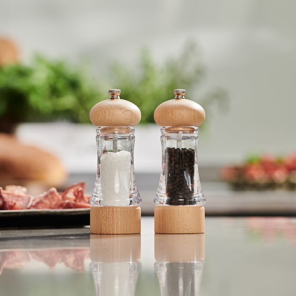 The Cole & Mason Basics Wood Salt & Pepper Mill Set features two clear mills with sturdy wood tops and bottoms, each with adjustable grind settings—one for coarse salt and one for whole black peppercorns—displayed side by side.
