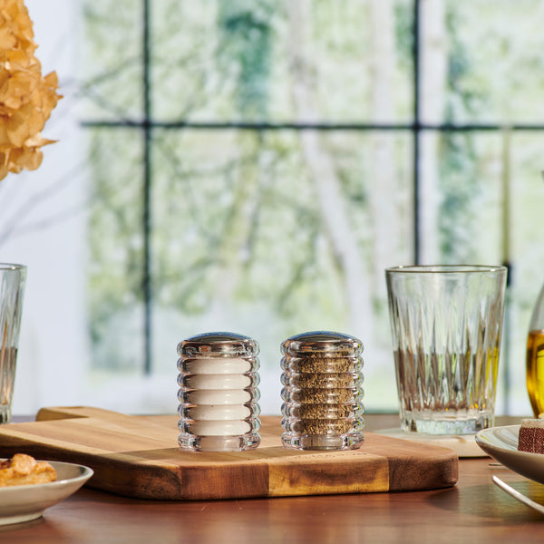 The Cole & Mason Beehive Shakers Set, featuring two durable acrylic shakers for salt and pepper, sits on a wooden board amid glasses and plates on a dining table, with blurred greenery visible through a window in the background.