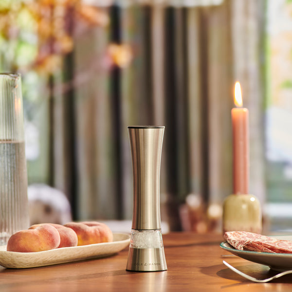 The Cole & Mason Burford Electronic Single Mill with adjustable grind settings stands on a wooden table beside peaches, a pink candle, water, and sliced meat, with a softly blurred natural light background.