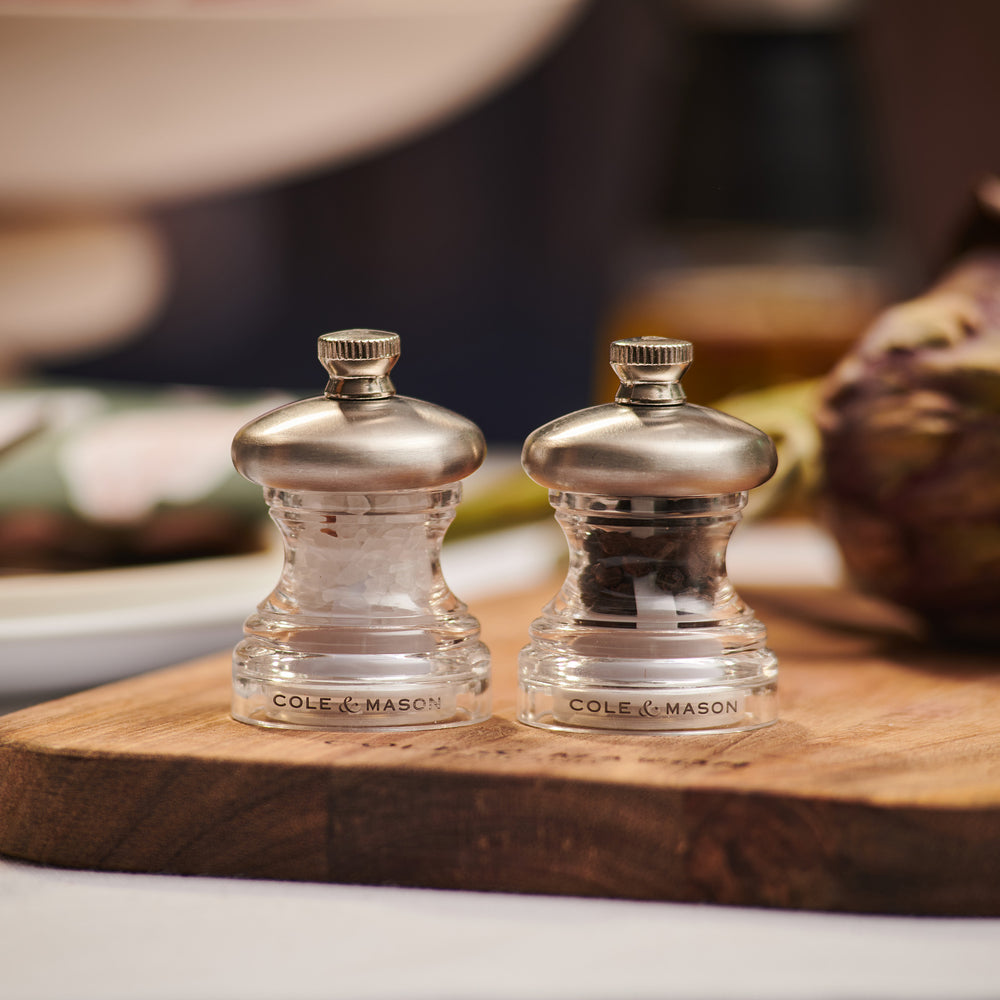 The Cole & Mason Button Salt & Pepper Mill Set features two clear grinders with metal tops labeled S and P. Compact and adjustable, one is filled with salt and the other with peppercorns, shown on a pale background.