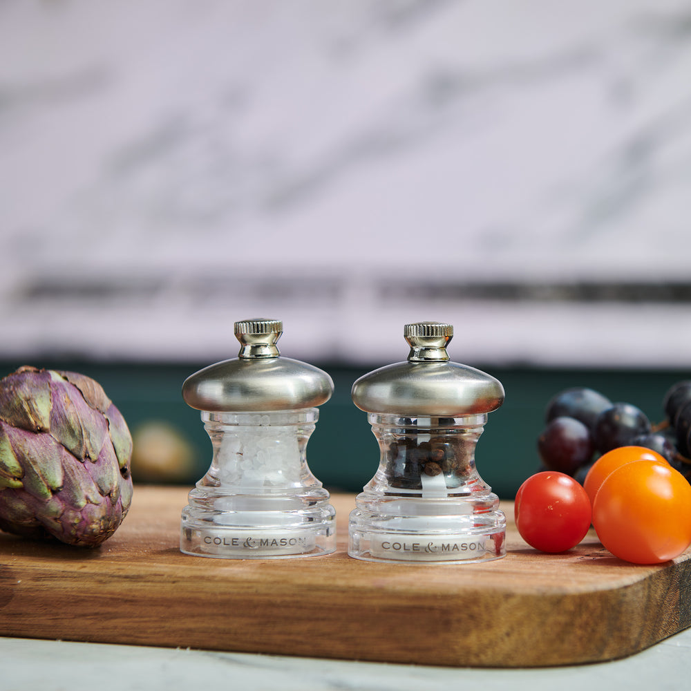 The Cole & Mason Button Salt & Pepper Mill Set features two clear grinders with metal tops labeled S and P. Compact and adjustable, one is filled with salt and the other with peppercorns, shown on a pale background.