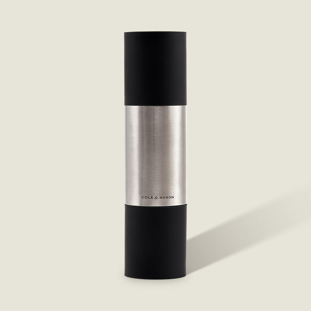 The Cole & Mason Clevedon 2-in-1 Mill features a modern cylindrical design with black matte ends and a brushed stainless steel center, standing upright on a light beige background—an innovative grinder ideal for contemporary kitchens.