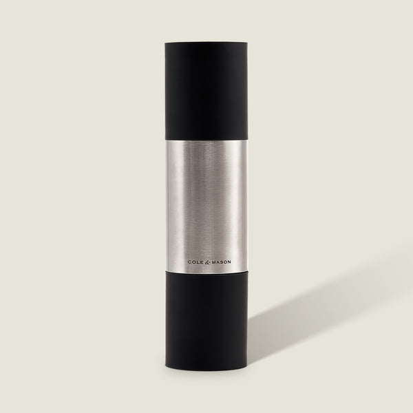 The Cole & Mason Clevedon 2-in-1 Mill features a modern cylindrical design with black matte ends and a brushed stainless steel center, standing upright on a light beige background—an innovative grinder ideal for contemporary kitchens.