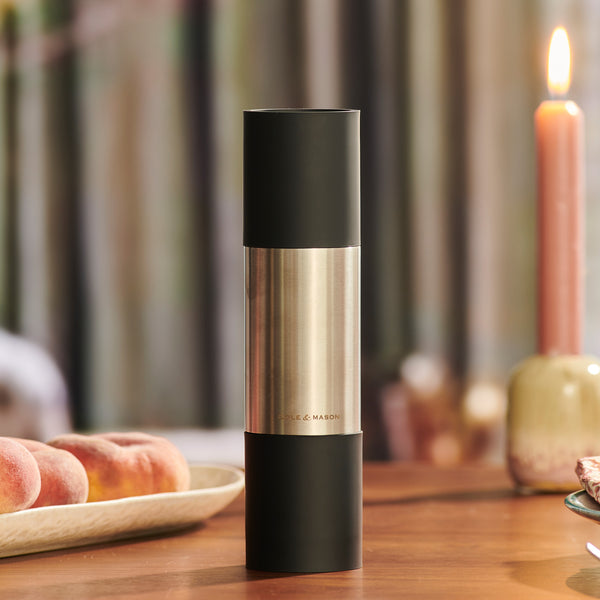 The Cole & Mason Clevedon 2-in-1 Mill, featuring a black top and bottom with a brushed metal center, sits on a wooden table beside peaches and a lit candle—ideal for modern kitchens seeking innovative grinders.