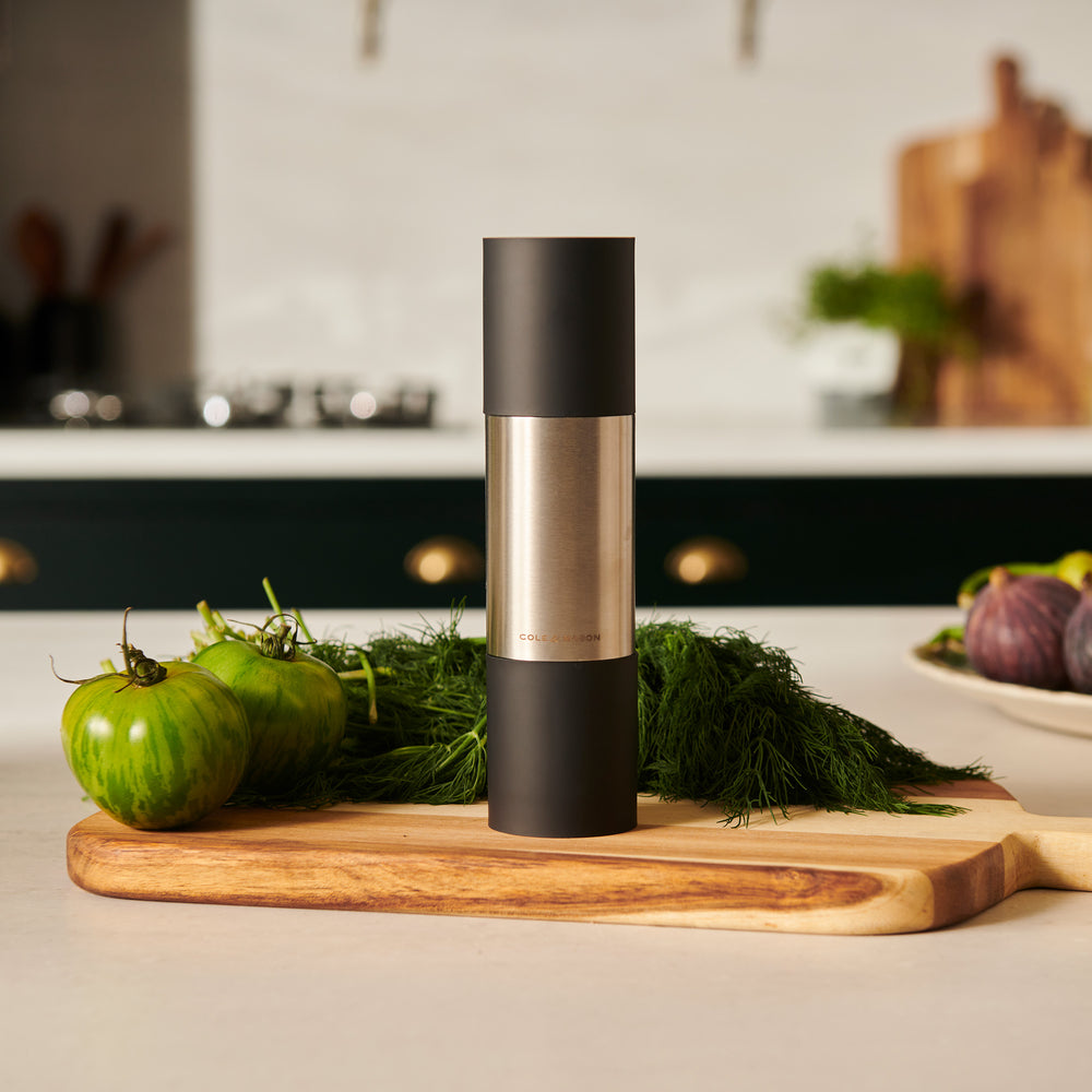 The Cole & Mason Clevedon 2-in-1 Mill features a modern cylindrical design with black matte ends and a brushed stainless steel center, standing upright on a light beige background—an innovative grinder ideal for contemporary kitchens.