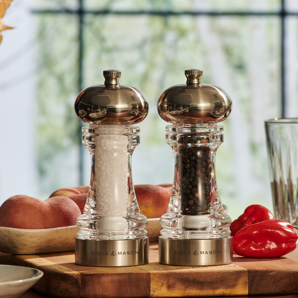 The Cole & Mason Clovelly Salt & Pepper Mill Set features two transparent mills with metallic tops—one containing white salt crystals, the other black peppercorns—both showcasing a modern design and adjustable grinding mechanisms on a light background.
