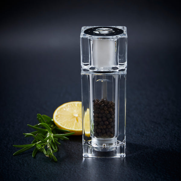 The Cole & Mason Cube Combi-Mill acrylic grinder, filled with black peppercorns, sits on a dark surface beside rosemary and lemon—a stylish modern kitchen accessory.