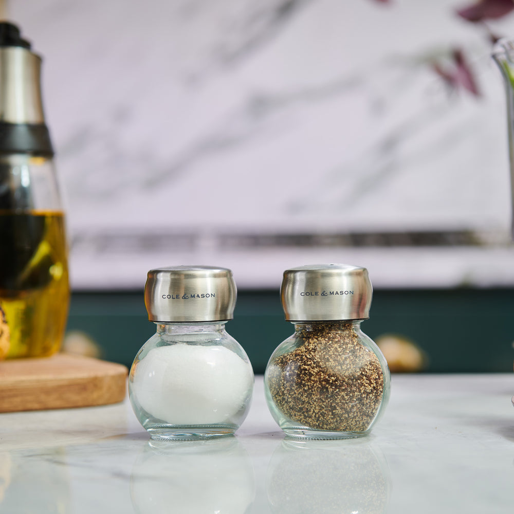 The Cole & Mason Darlington Shakers Set includes two glass jars with stainless steel lids—one empty, one filled with cracked black pepper. Both feature a sleek design and Cole & Mason branding, perfect for stylish seasoning.