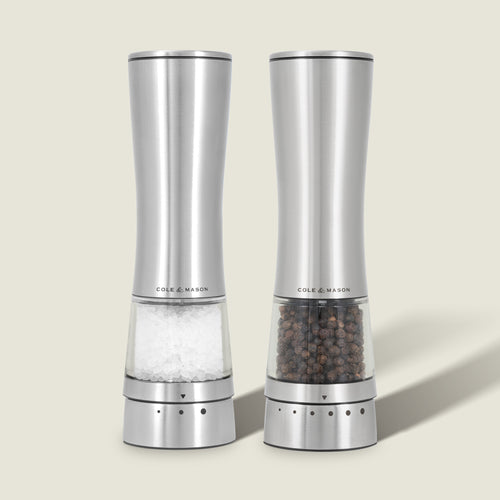 Two Cole & Mason Electric Derwent Salt & Pepper Mills stand side by side; one filled with coarse salt, the other with whole black peppercorns. Both feature stainless steel construction, clear bottoms, and adjustable grind settings.