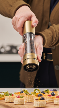 A person seasons small decorated pastry appetizers on a wooden board in the kitchen using Cole & Mason Derwent Gold Salt & Pepper Mills with advanced grinding technology.