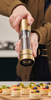 A person seasons small decorated pastry appetizers on a wooden board in the kitchen using Cole & Mason Derwent Gold Salt & Pepper Mills with advanced grinding technology.