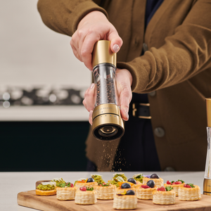 A person seasons small decorated pastry appetizers on a wooden board in the kitchen using Cole & Mason Derwent Gold Salt & Pepper Mills with advanced grinding technology.