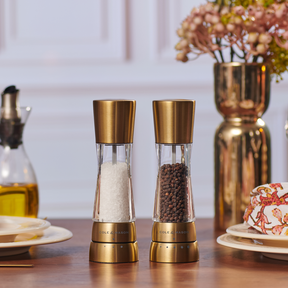 The Cole & Mason Derwent Gold Salt & Pepper Mills, filled with white salt crystals and black peppercorns, stand side by side on a neutral background, highlighting their advanced grinding technology.