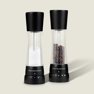 #salt&pepperset Two Cole & Mason Derwent Matt Black Salt & Pepper Mills, featuring adjustable grind settings and acrylic bodies, stand together on a light background, filled with coarse salt and whole black peppercorns.