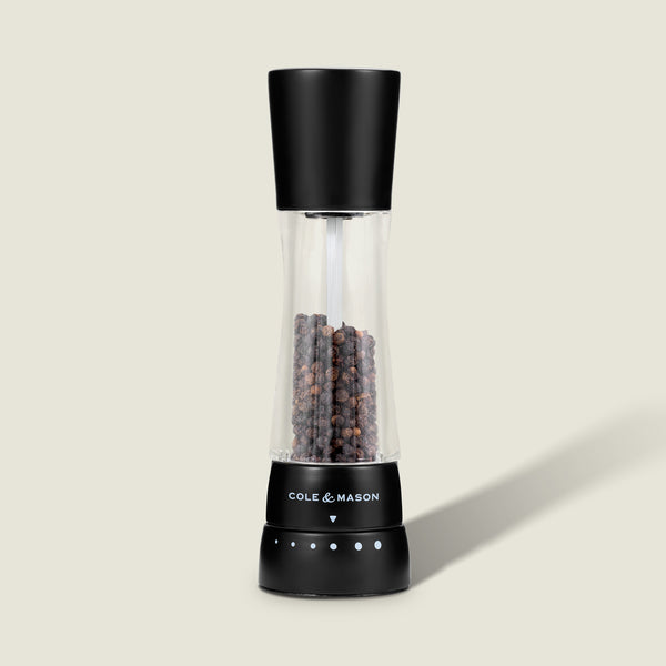 #pepper A Cole & Mason Derwent Matt Black Salt & Pepper Mill with an acrylic body and adjustable grind settings, filled with whole peppercorns, stands upright on a light beige surface with a subtle shadow to the right.
