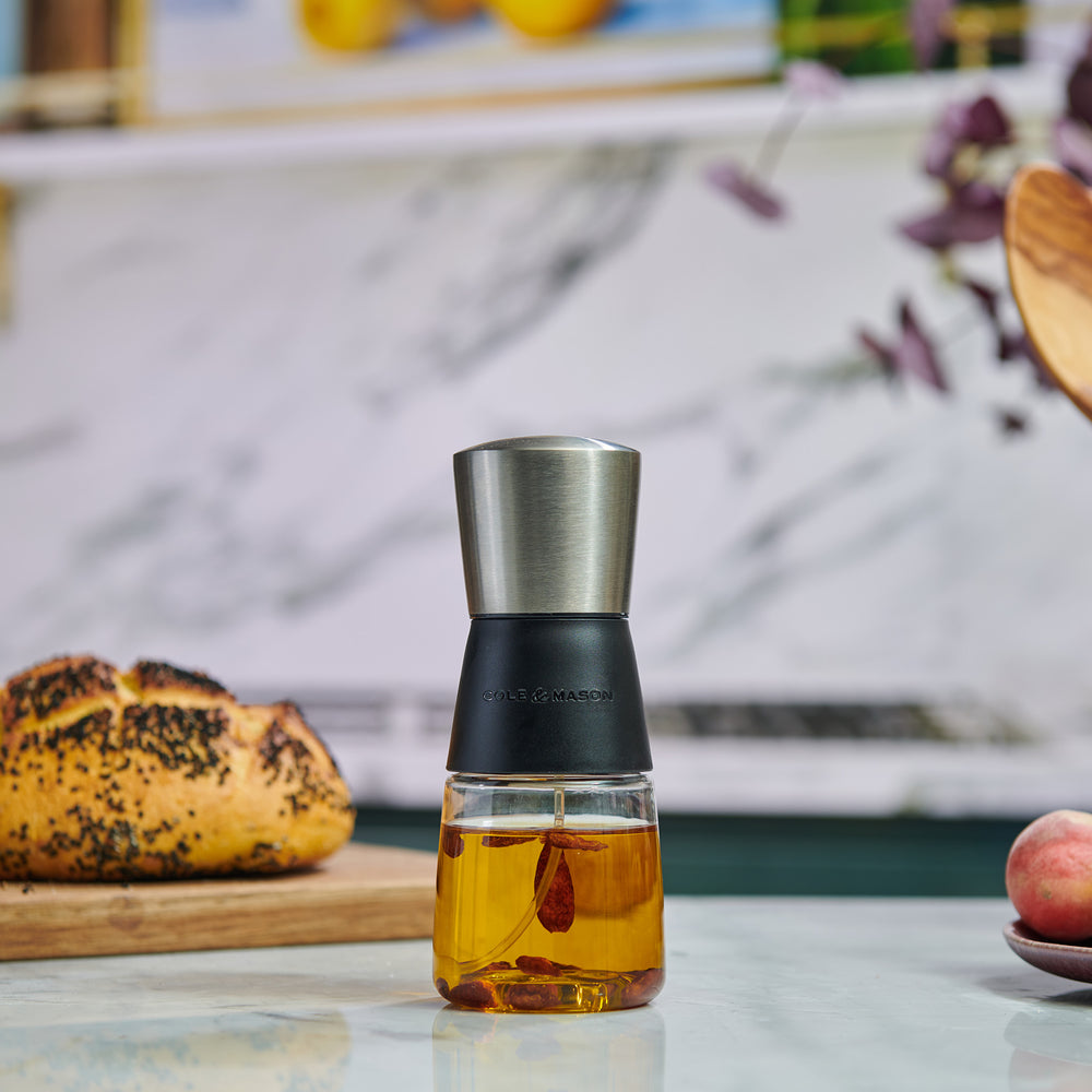 The Cole & Mason Epping Oil & Vinegar Mister features a stainless steel top, black middle section, and glass body filled with light yellow oil. Its fine misting mechanism helps promote healthier cooking, all set against a plain light background.