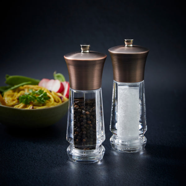 The Cole & Mason UK Exford Salt & Pepper Mill 160mm set, with precision grinders, sits on a dark surface with black peppercorns and white salt, a blurred dish behind—making it an ideal stylish gift.