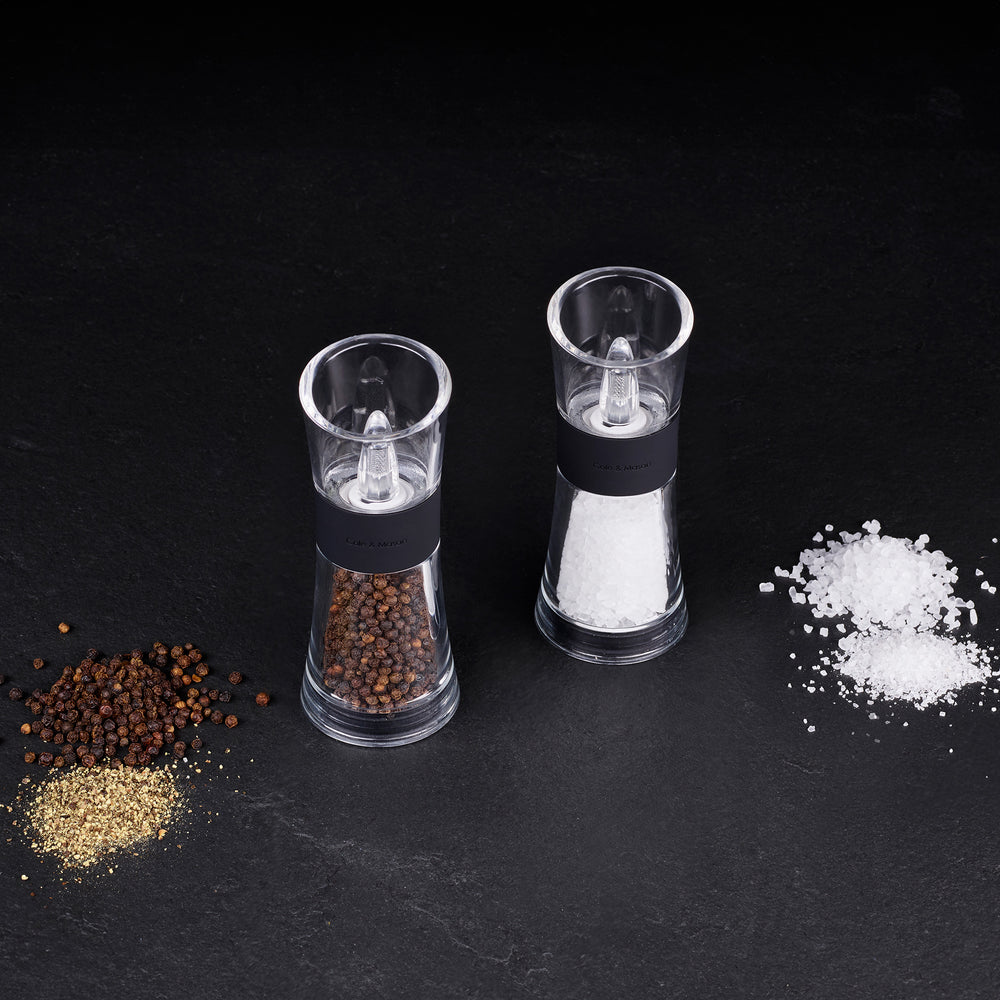 The Cole & Mason UK Flip Inverta Salt & Pepper Mill Set Black 155mm features two clear mills with black bands, one for coarse salt and one for whole black peppercorns, each with a quality grinding mechanism, shown on a light background.