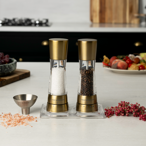 #gold The Cole & Mason Derwent Mill Set, Acrylic Tray & Funnel Set sits on a kitchen counter, surrounded by salt crystals, peppercorns, a metal funnel, and food items.