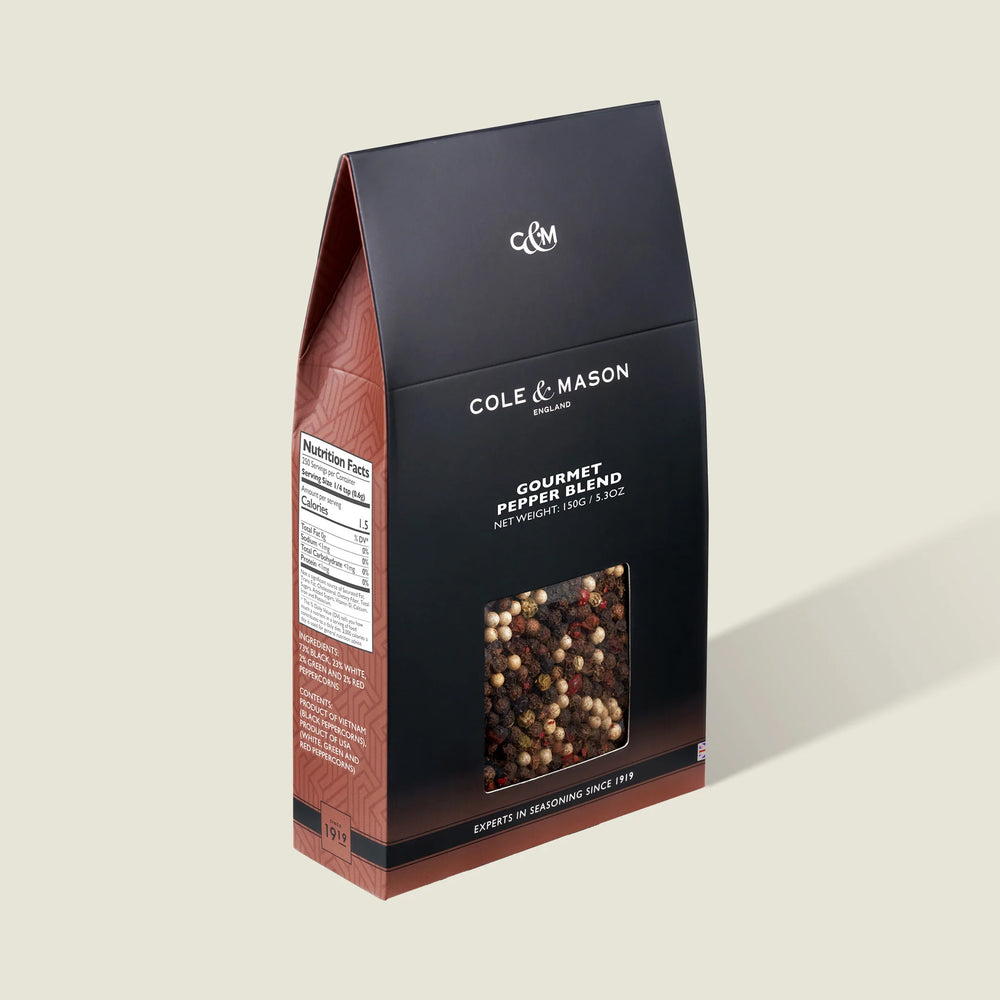 A box of Cole & Mason Gourmet Pepper Box features a clear window displaying mixed peppercorns. The mostly black packaging with brown accents is perfect for grinding fresh flavor and includes a nutrition facts label on the side.