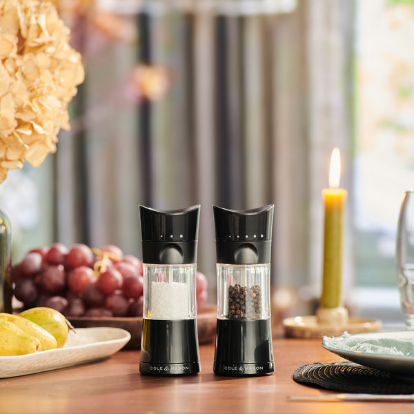 Two black Cole & Mason Harrogate Salt & Pepper Mills with adjustable grind settings add elegance and functionality to your meal, displayed on a wooden table amid grapes, pears, dried flowers, a lit candle, and plates.