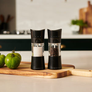 Two black and clear Cole & Mason Harrogate Salt & Pepper Mill Set grinders with adjustable grind settings rest on a wooden cutting board beside green tomatoes in a modern kitchen.